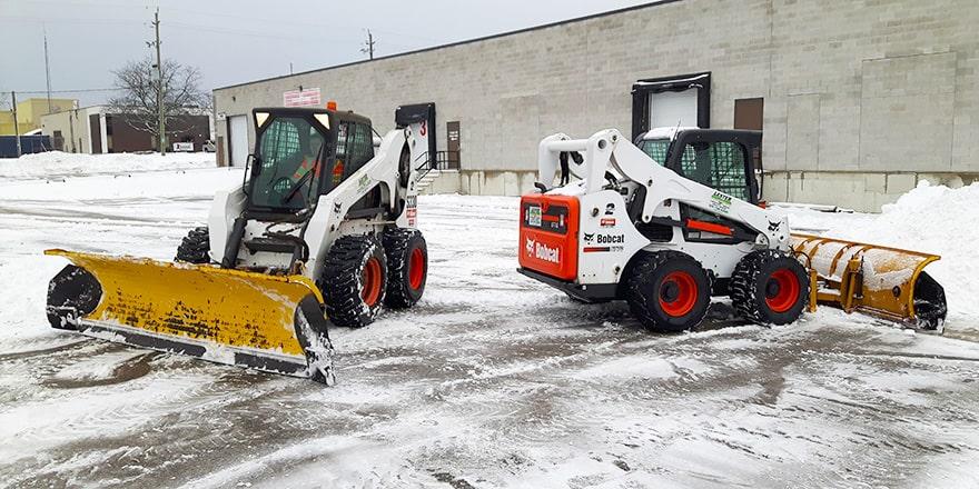 Skid steers with pusher blades clearing a commercial parking lot