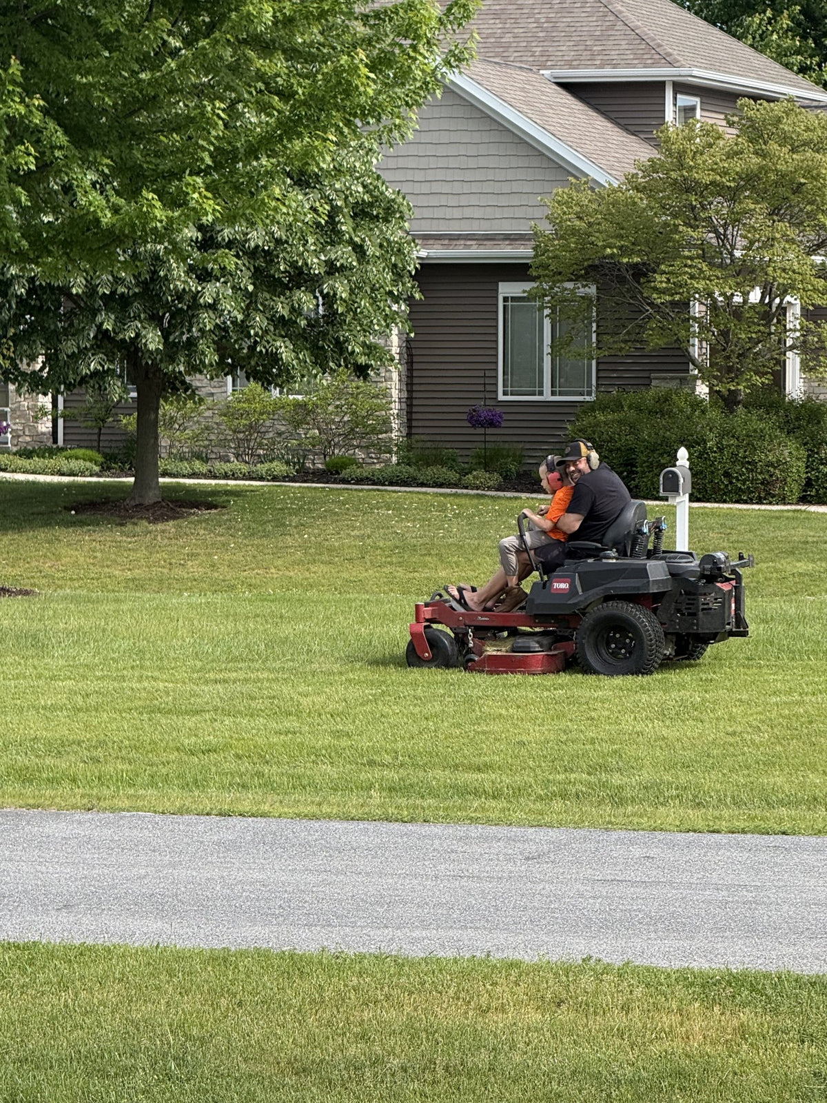 Mack and Carl Martens mowing together on the zero-turn — Mack on Carl's lap at the controls — for Sunny Stripes Landscaping in Sheboygan County