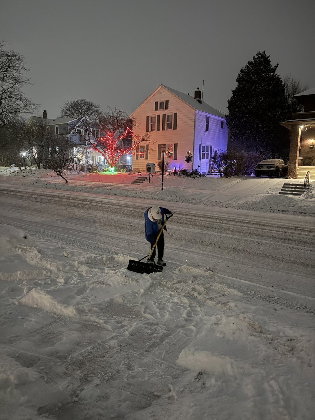 Mack clearing a sidewalk at night with a push shovel, Christmas lights on the houses behind him